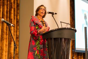 Speaker in colorful dress delivers presentation at podium against gold curtained background.