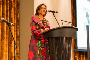 Presenter in floral dress speaking at podium during conference event with projection screen visible.