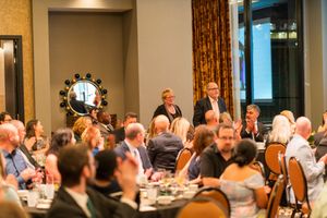 Crowded formal dinner event with guests seated at round tables in an elegant room with ornate curtains.