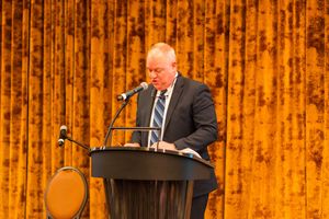 Speaker presenting at a podium against a golden curtain backdrop.