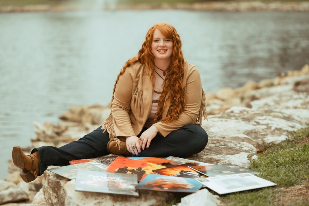 A student sits by a lake studying with textbooks and papers spread out on the grass in an autumn setting.
