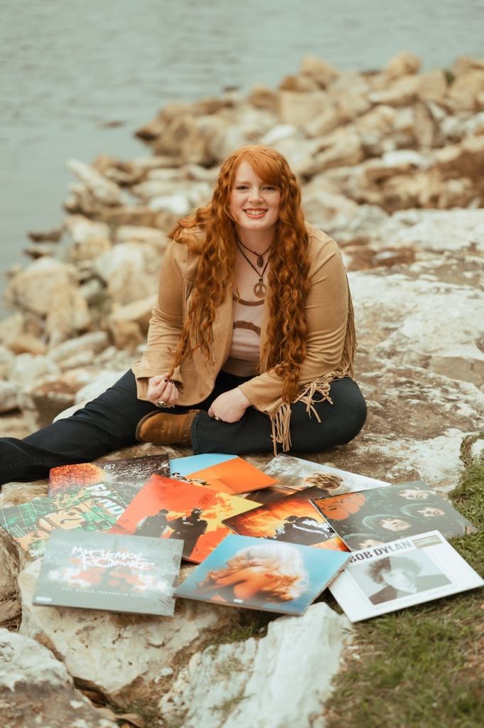 A person sits on a rocky shore with scattered vinyl records displayed around them in a casual outdoor setting.