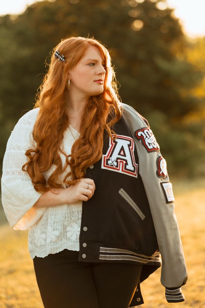 A person in a white lace top and varsity jacket poses against a golden sunset backdrop.