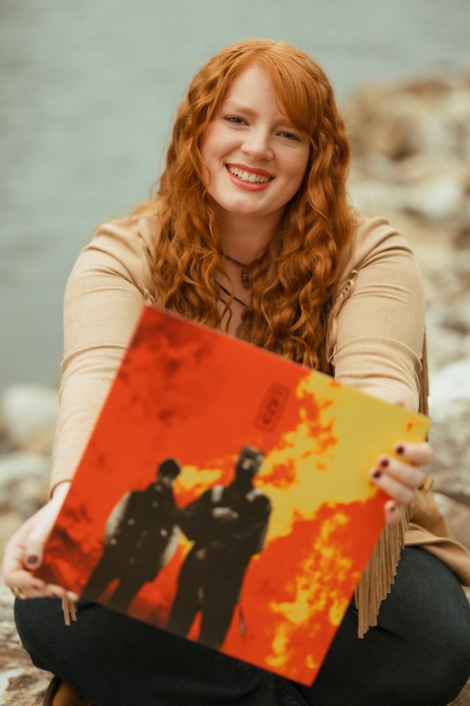 Woman holds vintage record album with orange and yellow cover while sitting outdoors.