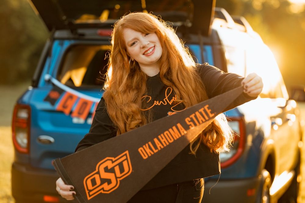 A person holds an Oklahoma State University pennant in front of a blue car during golden hour.