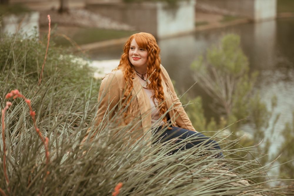 Red-haired figure sits outdoors near a lake surrounded by autumn foliage and brush.