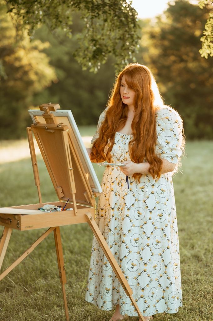 A person with long red hair paints at an easel outdoors wearing a floral dress at sunset.