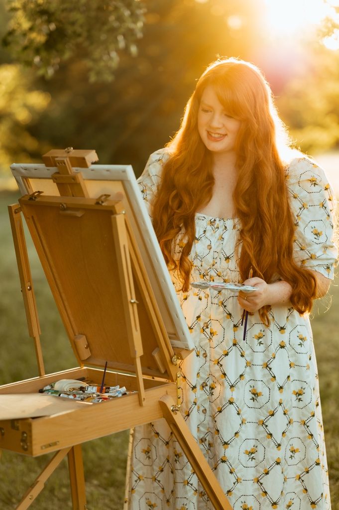 Artist in floral dress works at wooden easel during golden sunset hour.