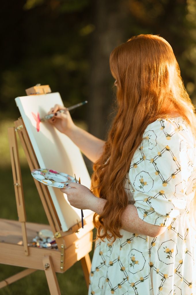 A person in a floral dress paints on a canvas at an outdoor wooden easel.