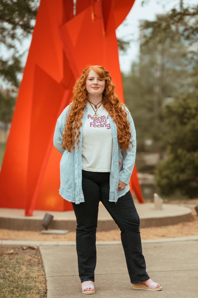 Woman with flowing red hair stands in front of large red outdoor sculpture wearing casual attire.