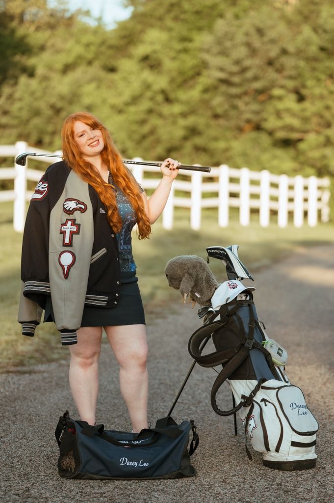 A golfer stands by a golf bag and cart on a path with a white fence in the background.