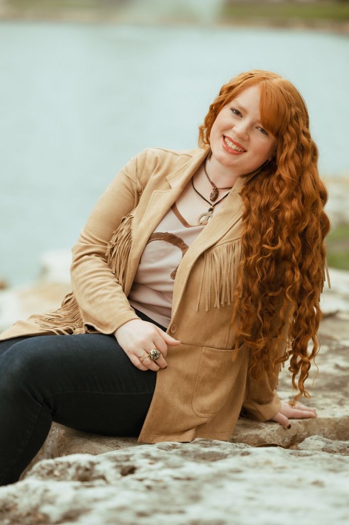 Woman with long wavy red hair wearing tan fringe jacket poses on rocky shoreline near water.