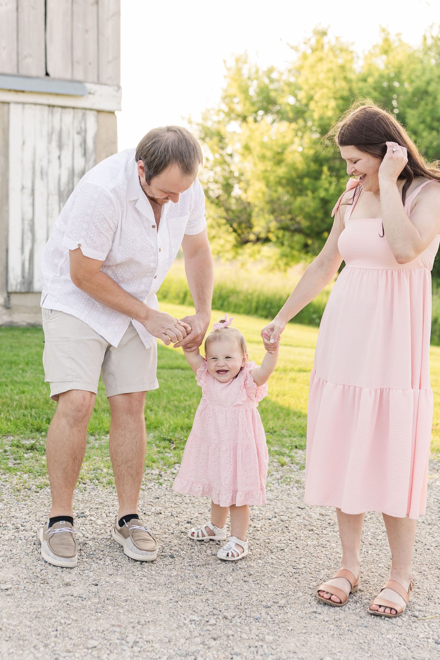 A happy family in coordinating white and pink outfits holds hands and plays together on a dirt path near a rustic barn.