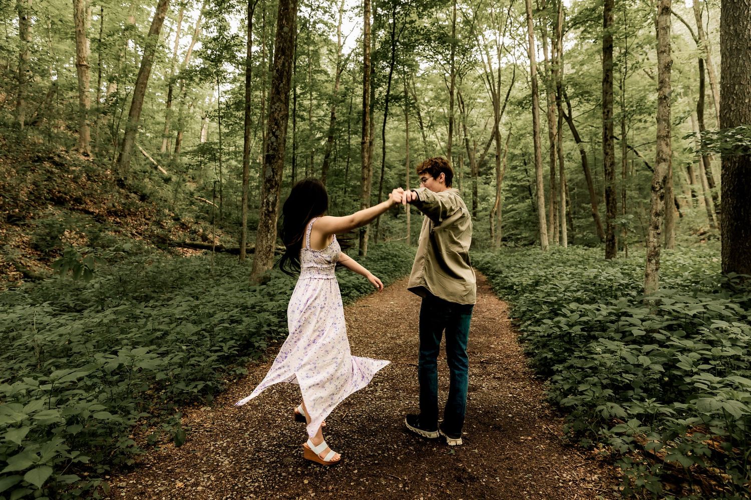 Two people dance together while holding hands on a forest path surrounded by lush green foliage.