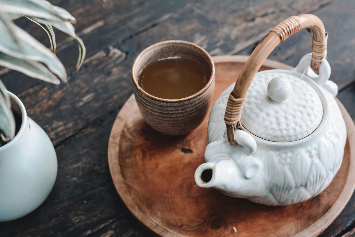 White ceramic teapot and clay cup on wooden tray with houseplant in background.