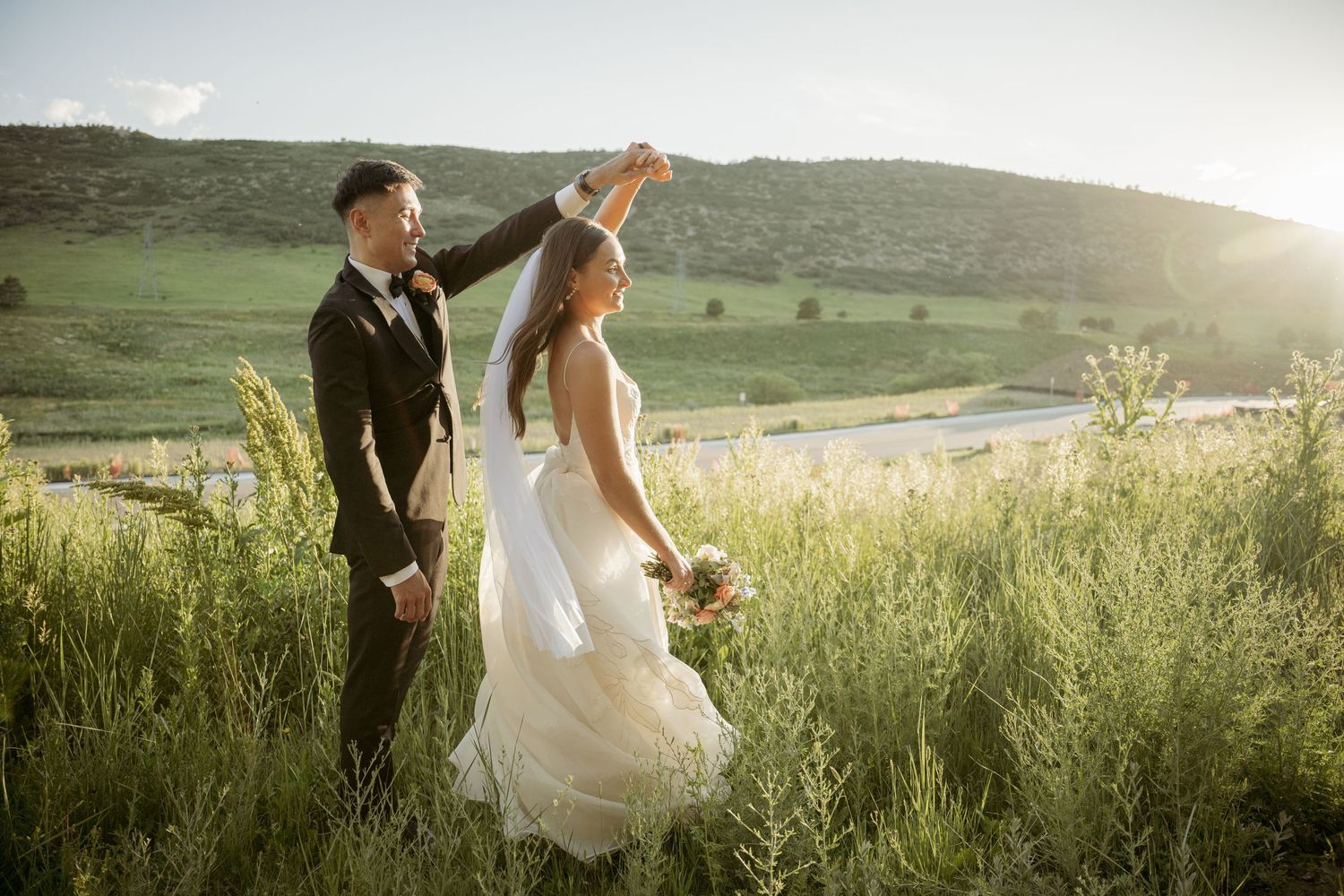 A couple in wedding attire holding hands while walking through a sunlit forest on a hillside.
