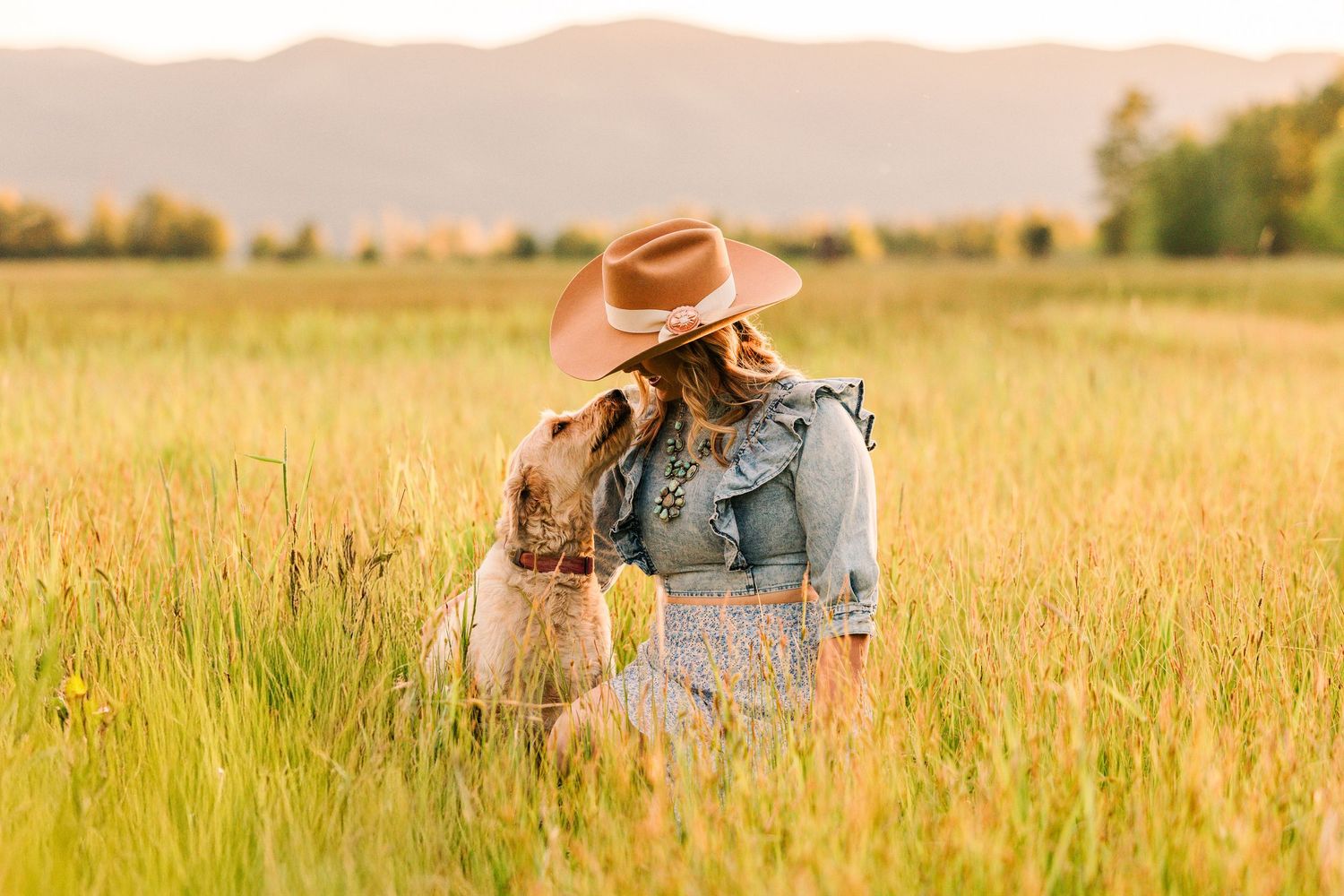 Person in denim and brown hat sitting in golden wheat field with dog during sunset with mountains in background.