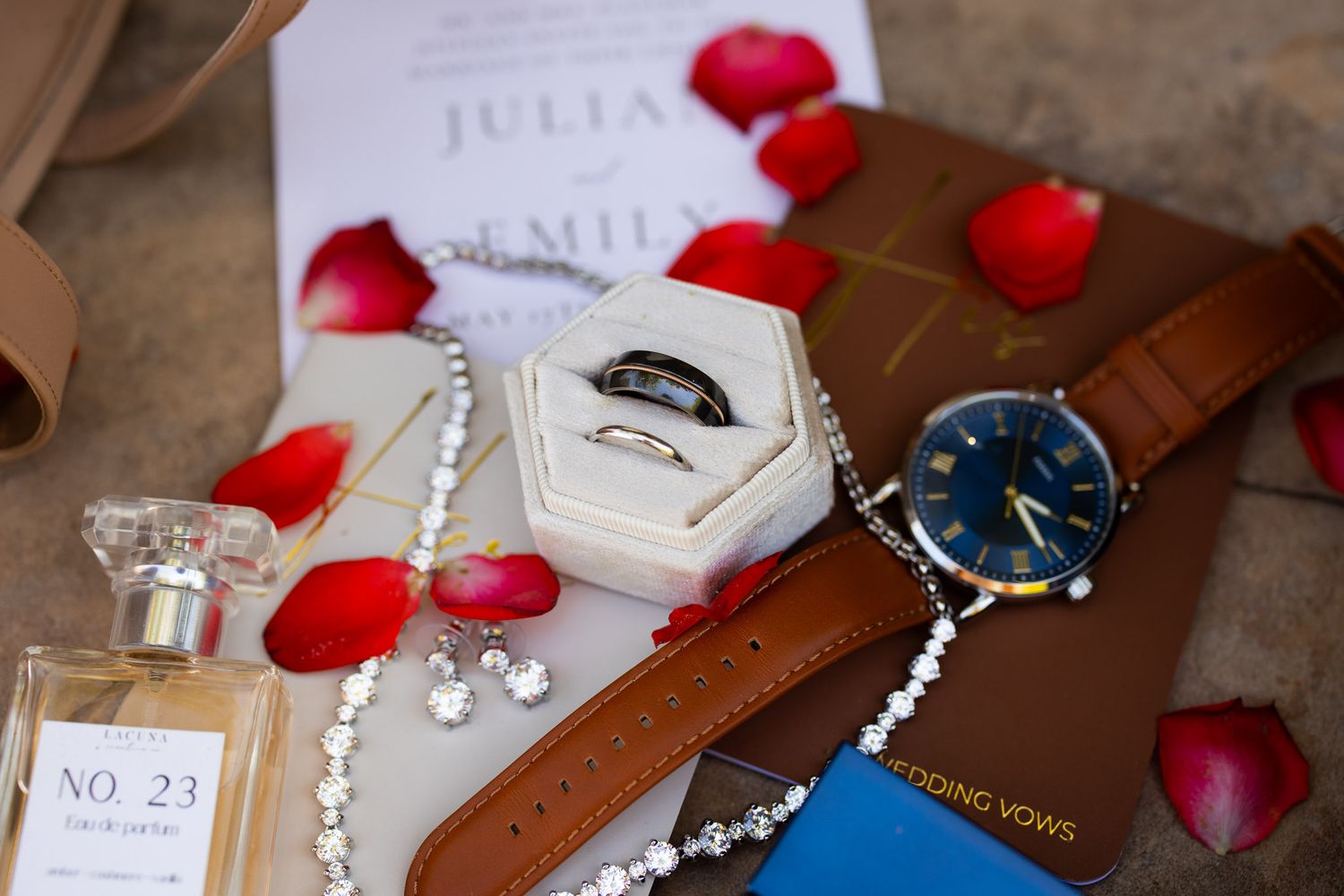 A luxury flat lay with engagement ring box, leather watch, pearl necklace and scattered red rose petals on a textured surface.