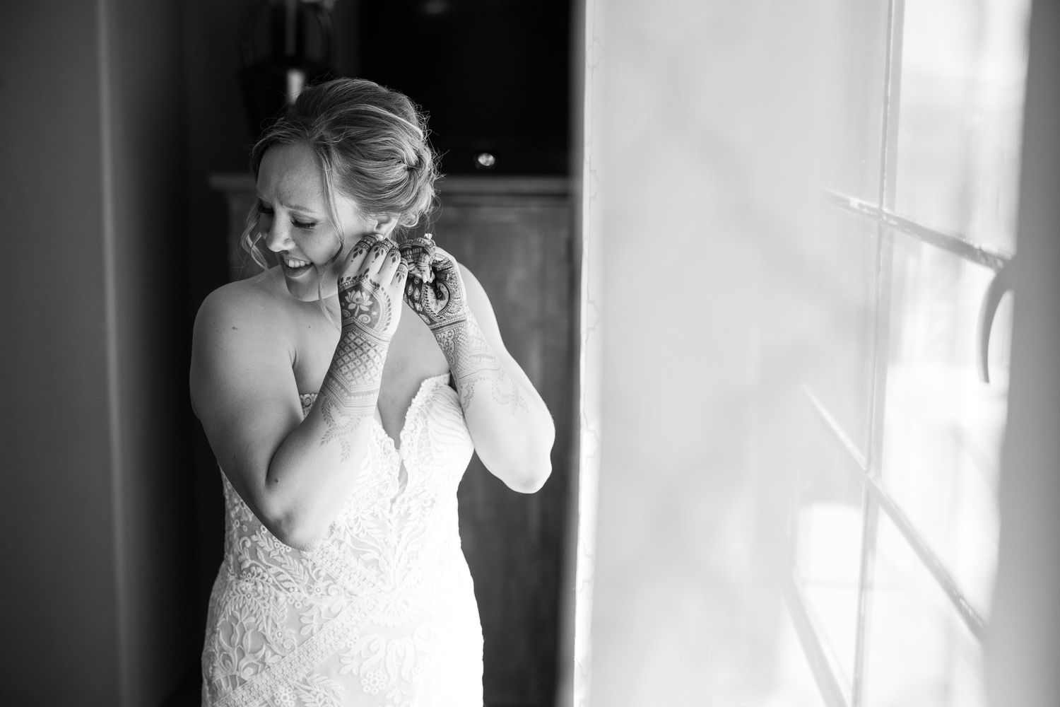 Black and white photo of bride putting on earrings by window light.