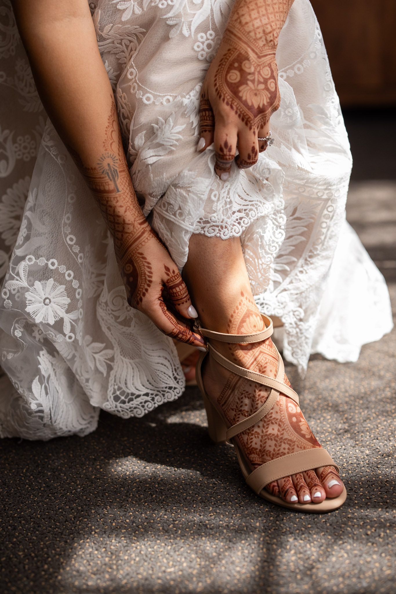 Bride wearing white lace dress adjusts strappy sandal showing intricate henna designs on hands and feet.