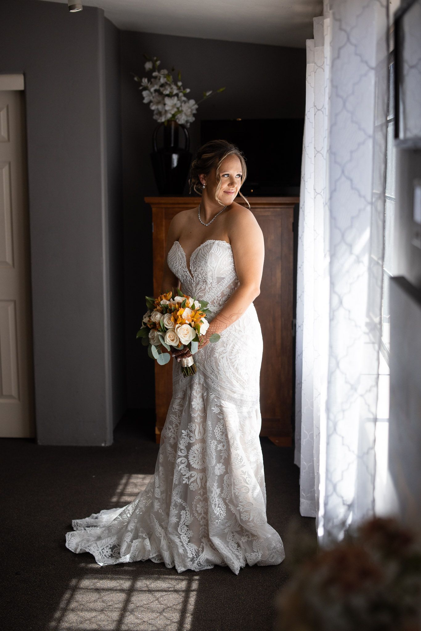 Bride in lace wedding dress holding autumn bouquet poses by window.