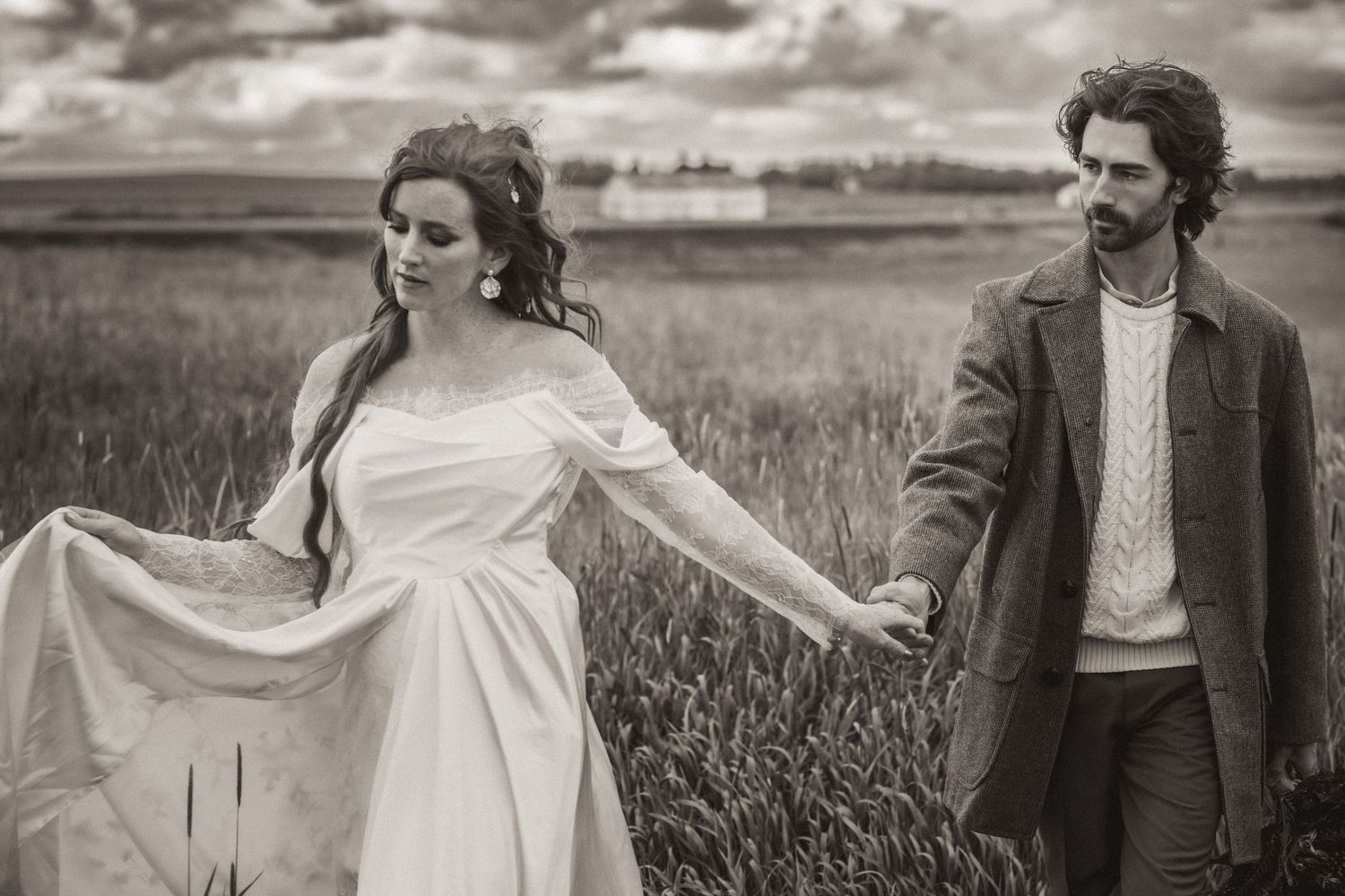 A couple walks together through a scenic wheat field on a cloudy day with the person in white spinning playfully.