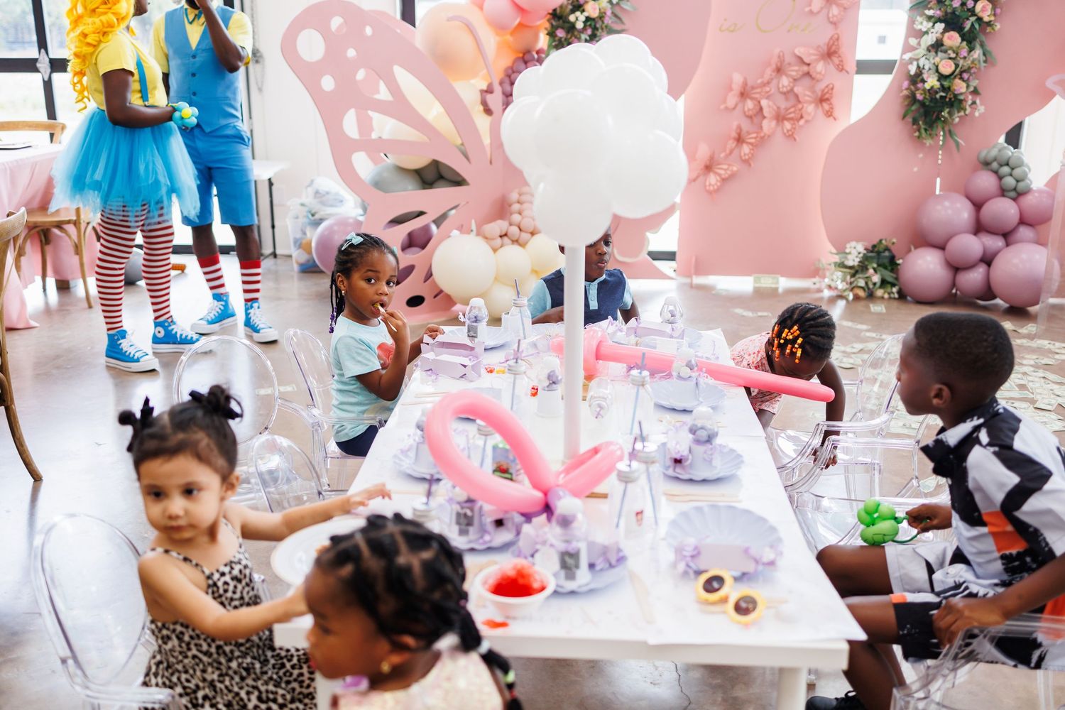Children gather around a decorated pink and white table at a birthday party with balloons and party decorations.