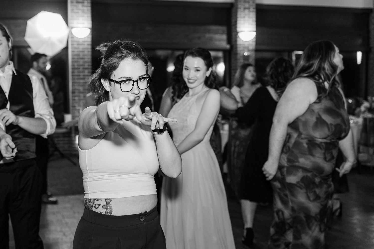 A partygoer dances while taking a sip from a drink at a wedding reception as other guests celebrate in the background.