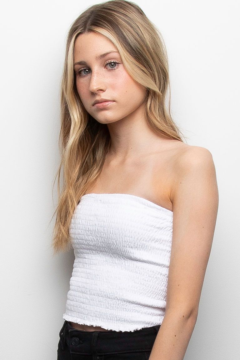 Young model poses against white background wearing strapless white smocked top and dark pants with blonde hair.