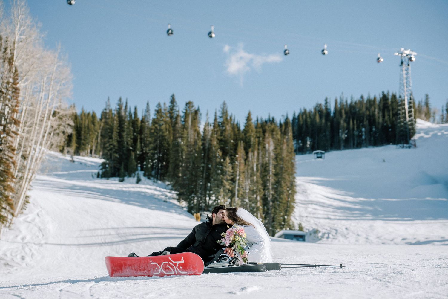 Skier resting in snow on sunny winter day with evergreen trees and ski slopes in the background.