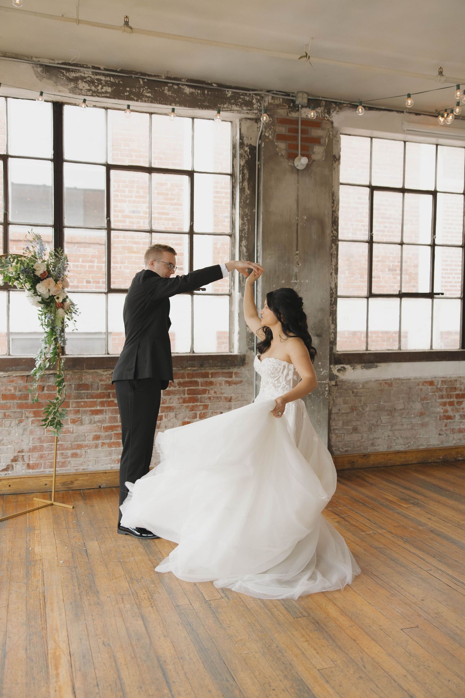 Wedding couple dances gracefully in a rustic industrial loft space with exposed brick walls and large windows.