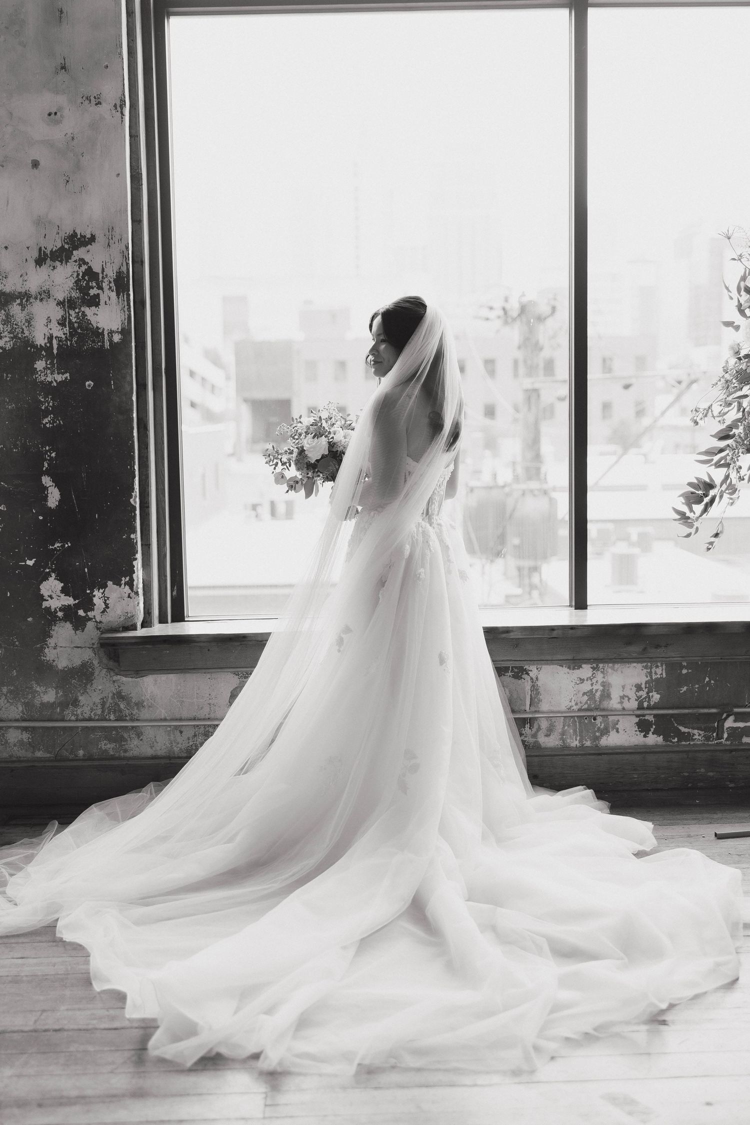 Elegant black and white photograph of a bride in a flowing wedding gown with cathedral veil gazing out a large window.