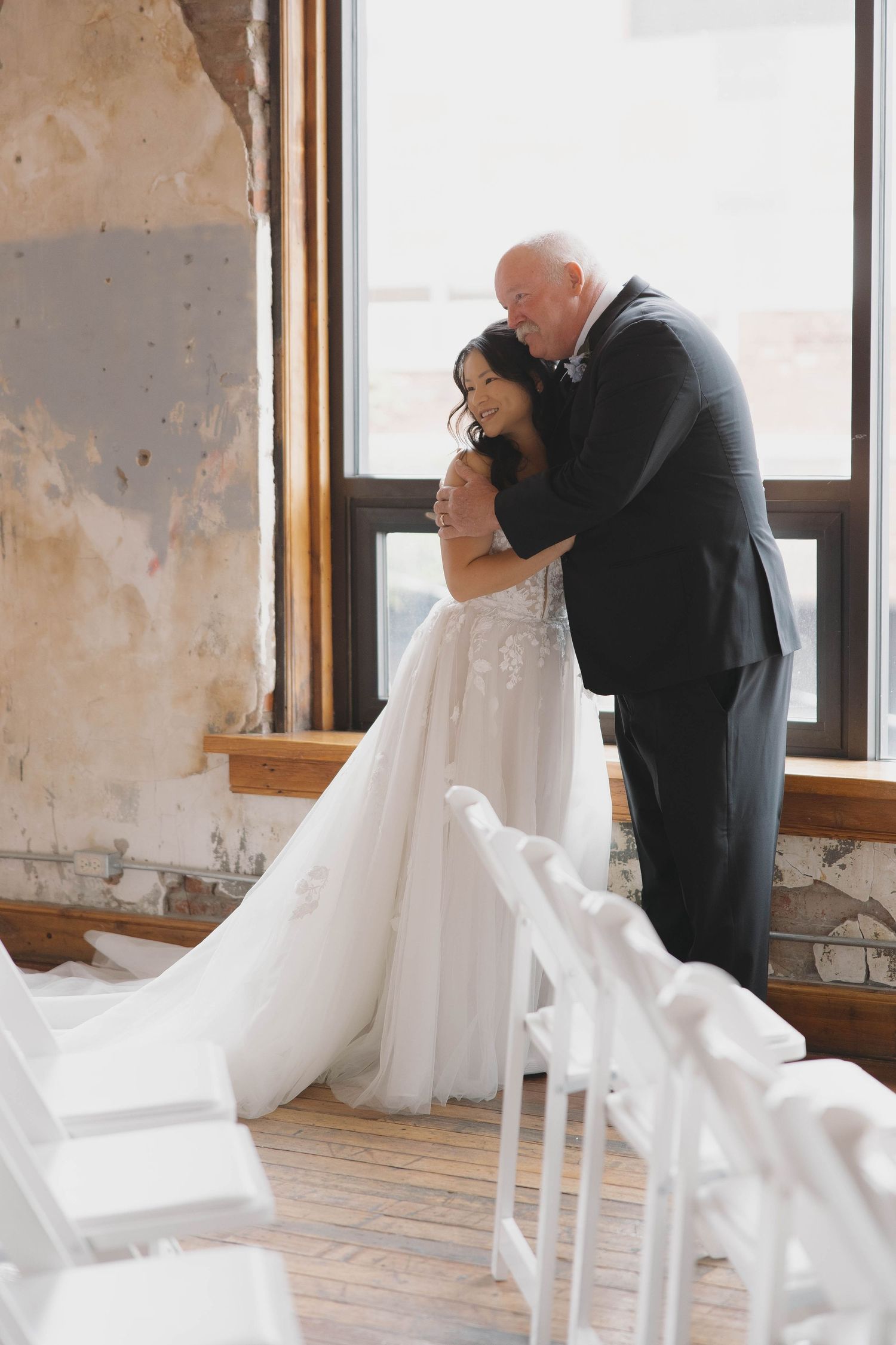 A tender romantic moment of a couple embracing in a rustic room with large windows and a flowing white wedding gown.