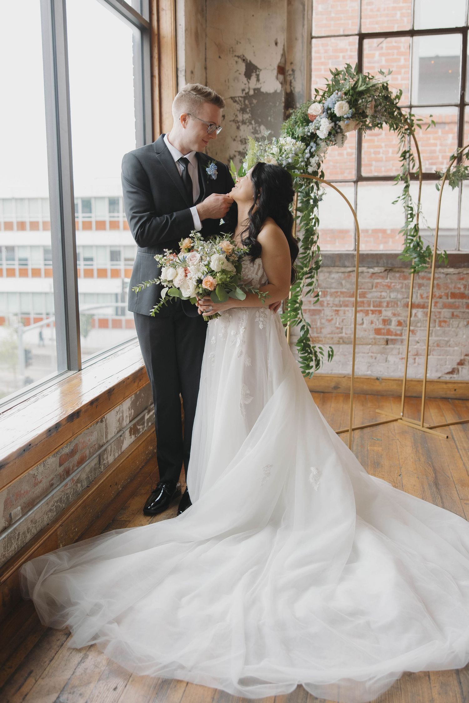 A couple shares an intimate moment in front of an elegant floral arch by large windows in an industrial-style wedding venue.
