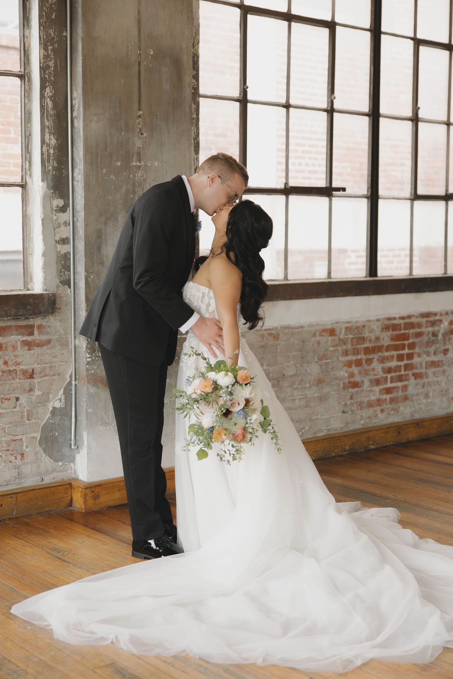 Elegant bride in flowing white dress holds a pastel bouquet as couple shares romantic moment in rustic industrial venue.