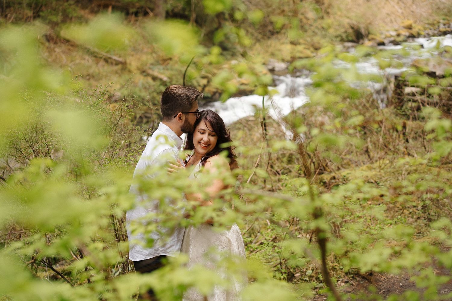 A couple embraces on a hillside with a flowing stream in the background, viewed through blurred foliage.