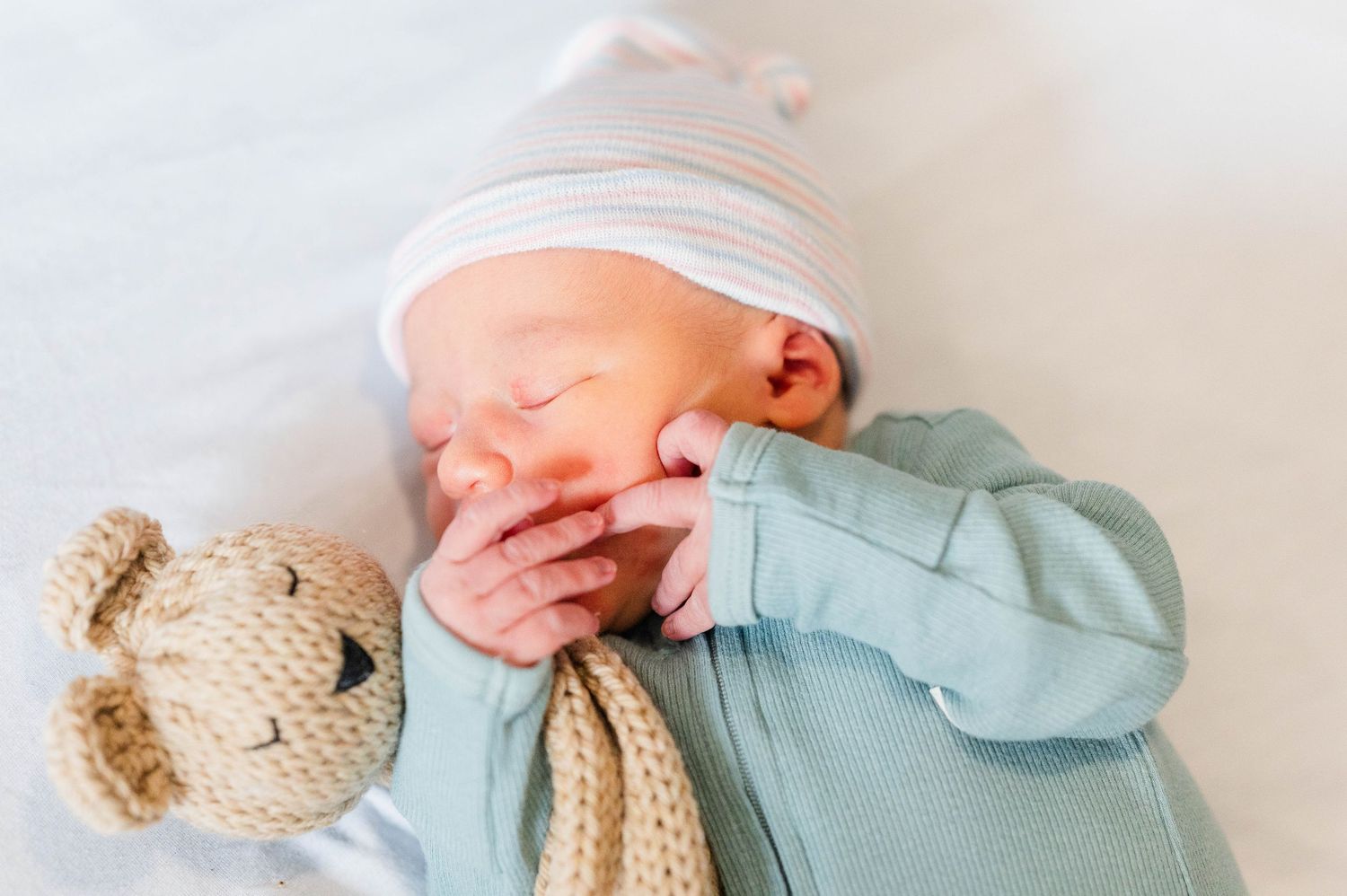 Newborn baby in a mint green onesie yawns next to a knitted teddy bear while wearing a white hat.