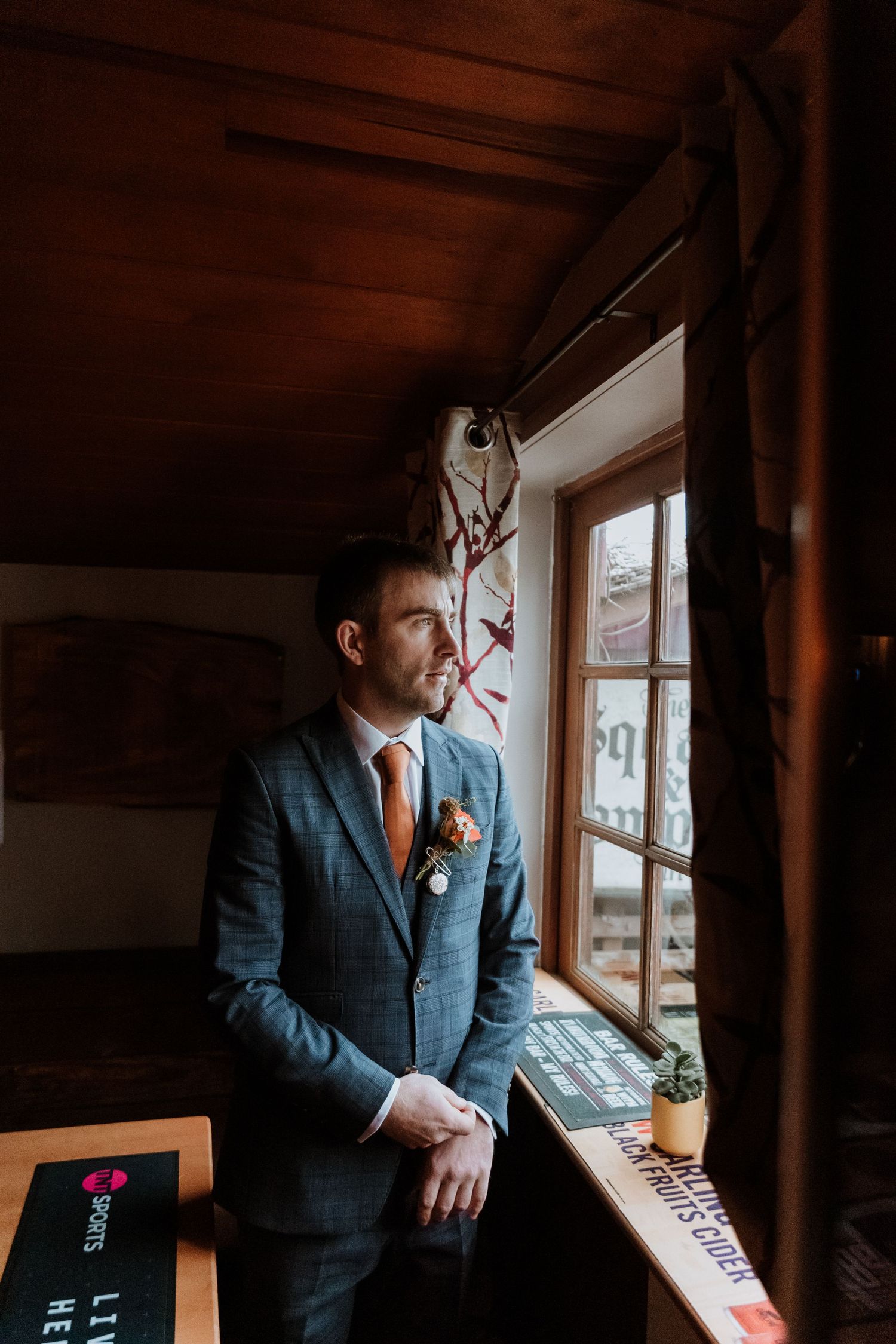 Groom in a blue suit stands by a rustic window, with warm interior lighting casting a moody atmosphere near Forden.