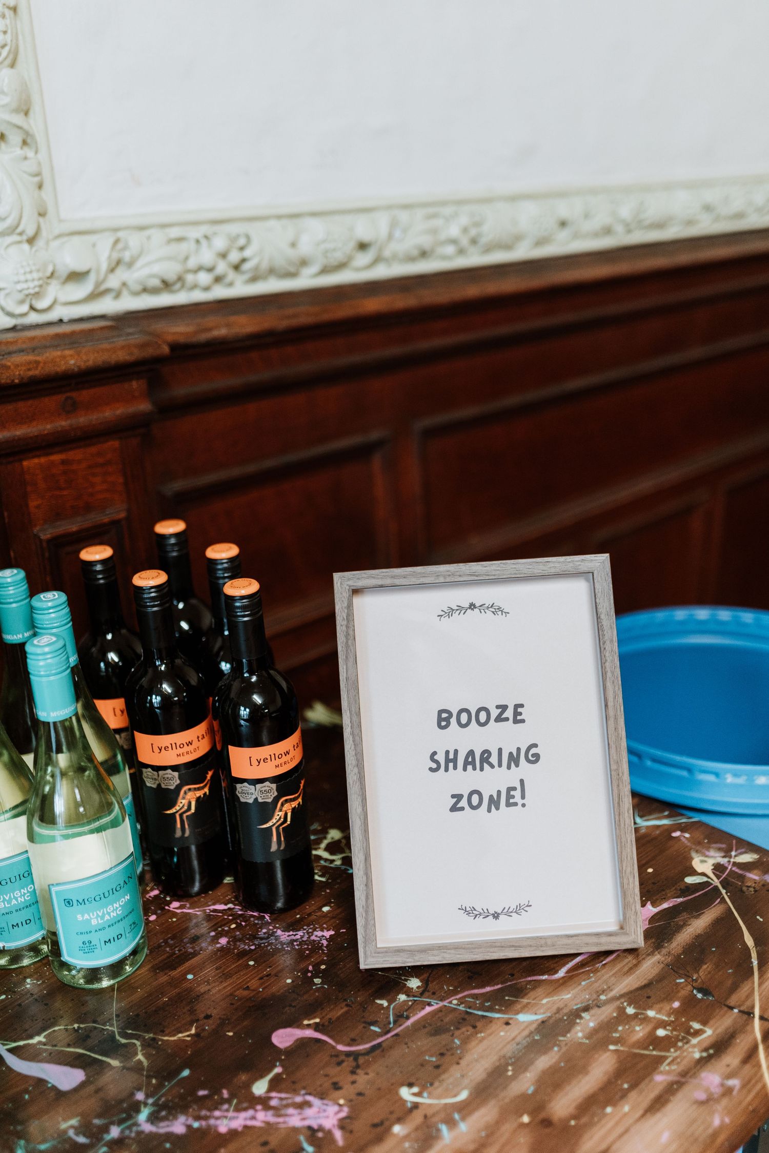 A framed 'Booze Shaking Zone' sign displayed on a wooden table next to wine bottles and turquoise glassware.