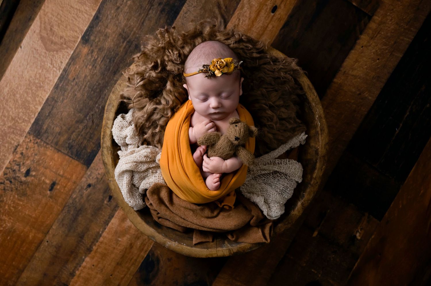 A rustic newborn portrait in a wooden bowl with orange and beige wrapping on wooden floor.