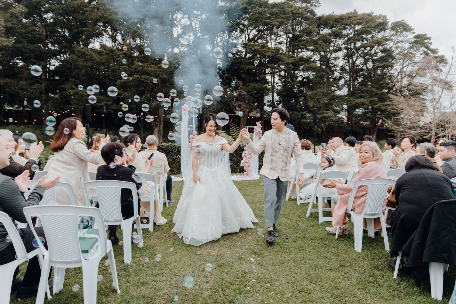Bubbles float through the air as wedding guests celebrate on a grassy lawn with white chairs.