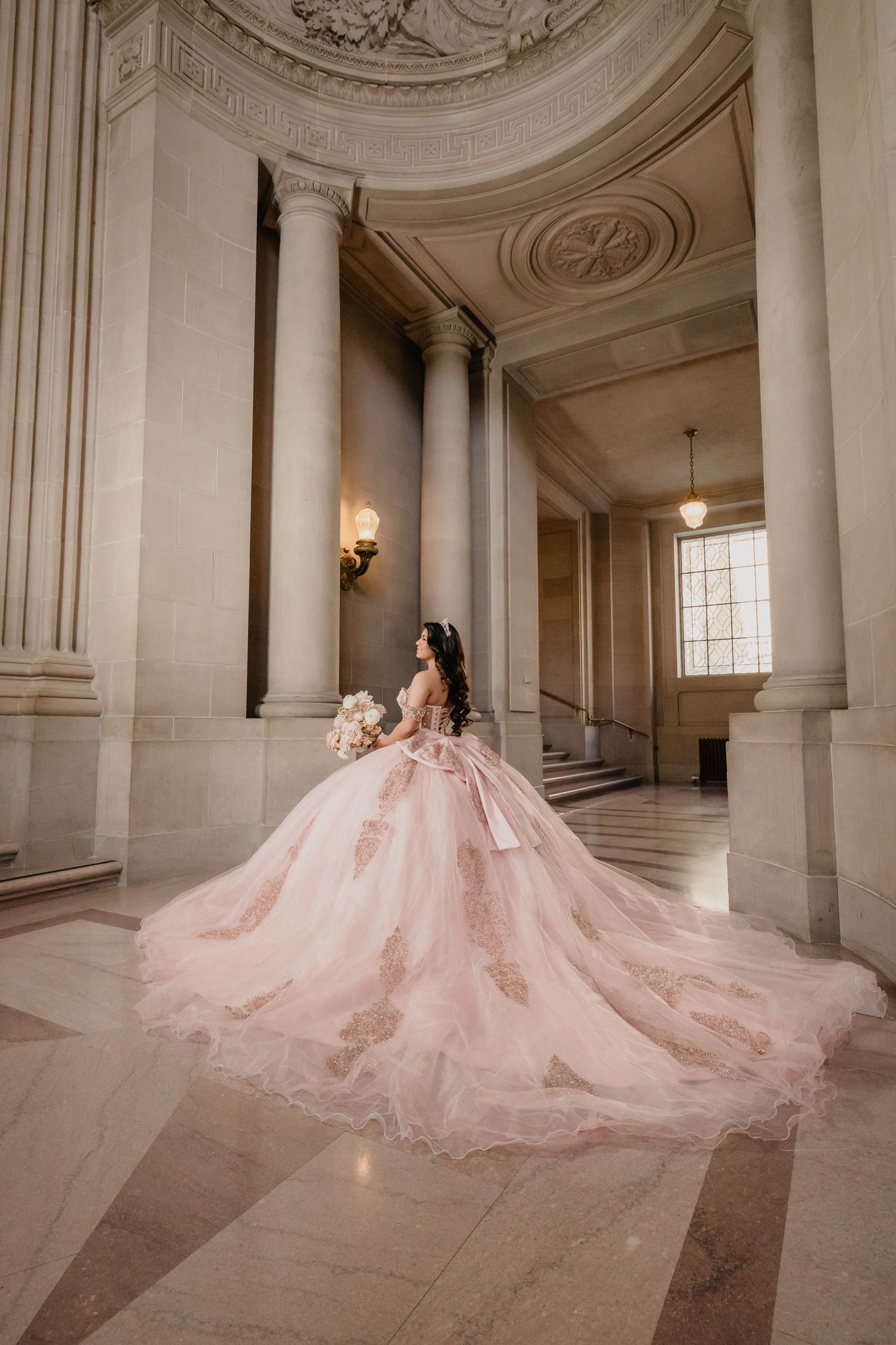 A quinceanera in a blush pink ball gown with a dramatic train poses in an elegant marble hallway with classical columns.