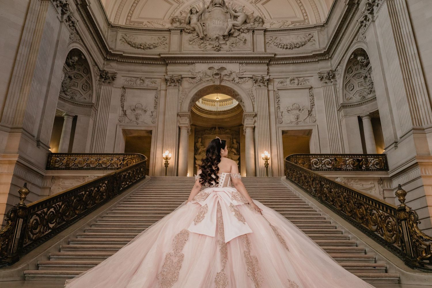 Stunning pink princess quinceanera captured from behind on elegant marble staircase with ornate architectural details.