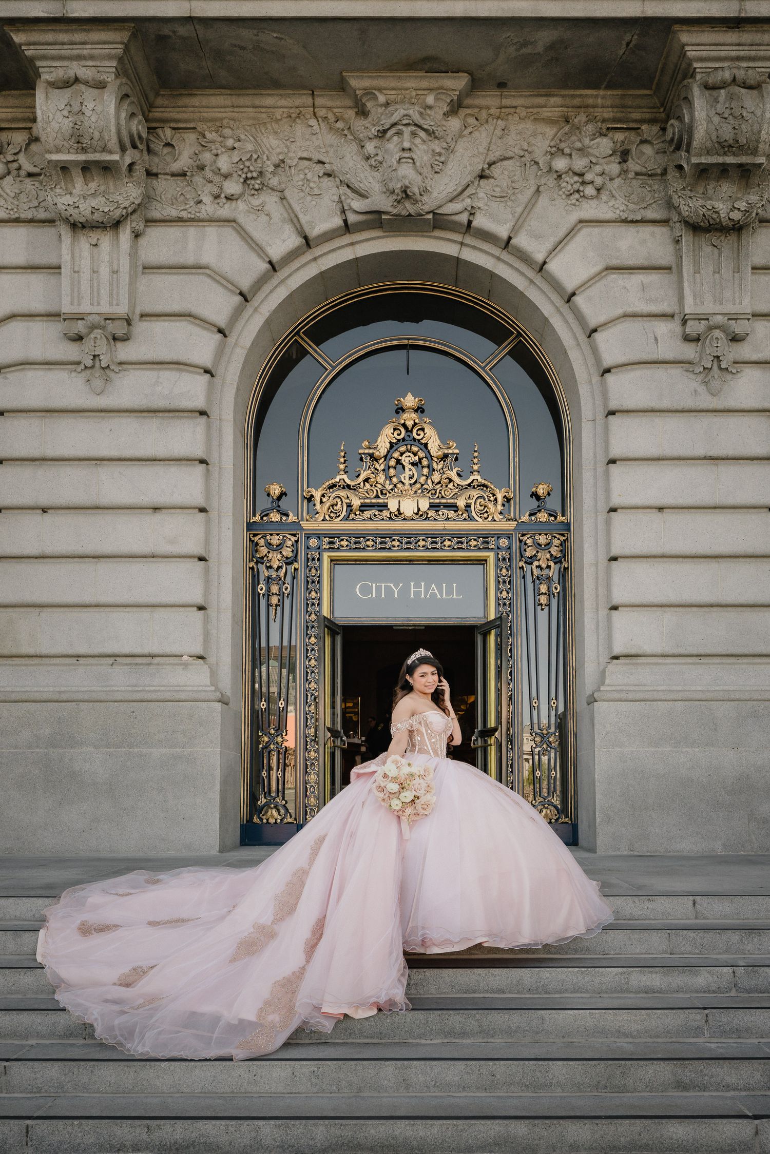 A flowing blush pink quinceanera with long train photographed on the steps of an ornate City Hall entrance.