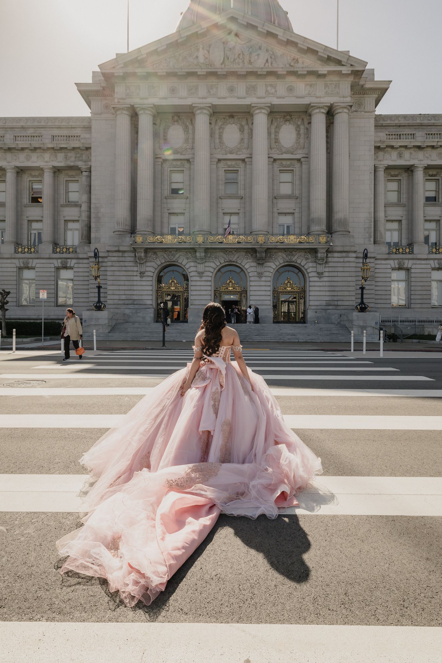 A Quinceanera in a flowing pink dress walks in front of San Francisco City Hall on a sunny day.