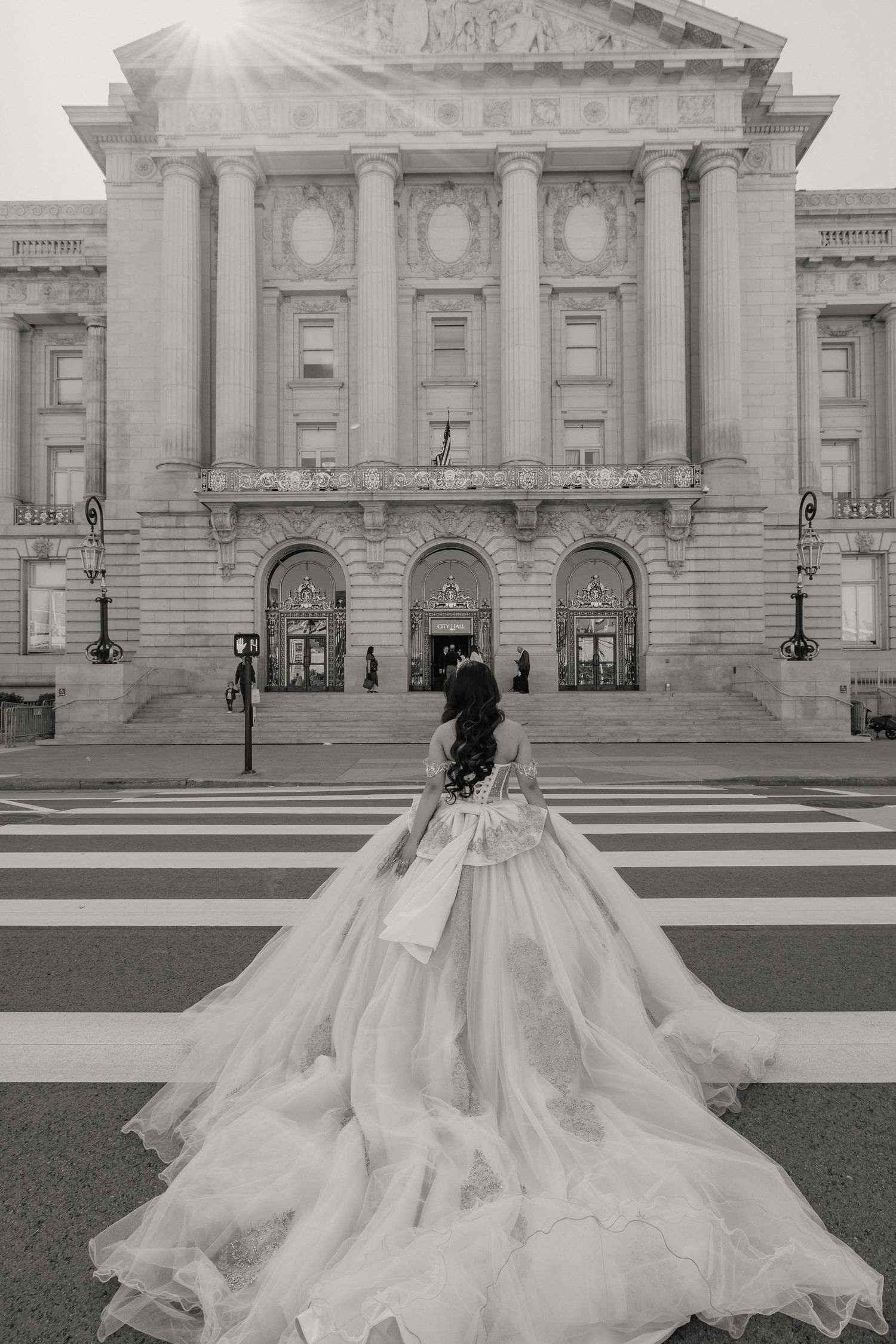 Black and white photo of a quinceanera in a flowing ballgown dress standing on steps of a grand neoclassical building.