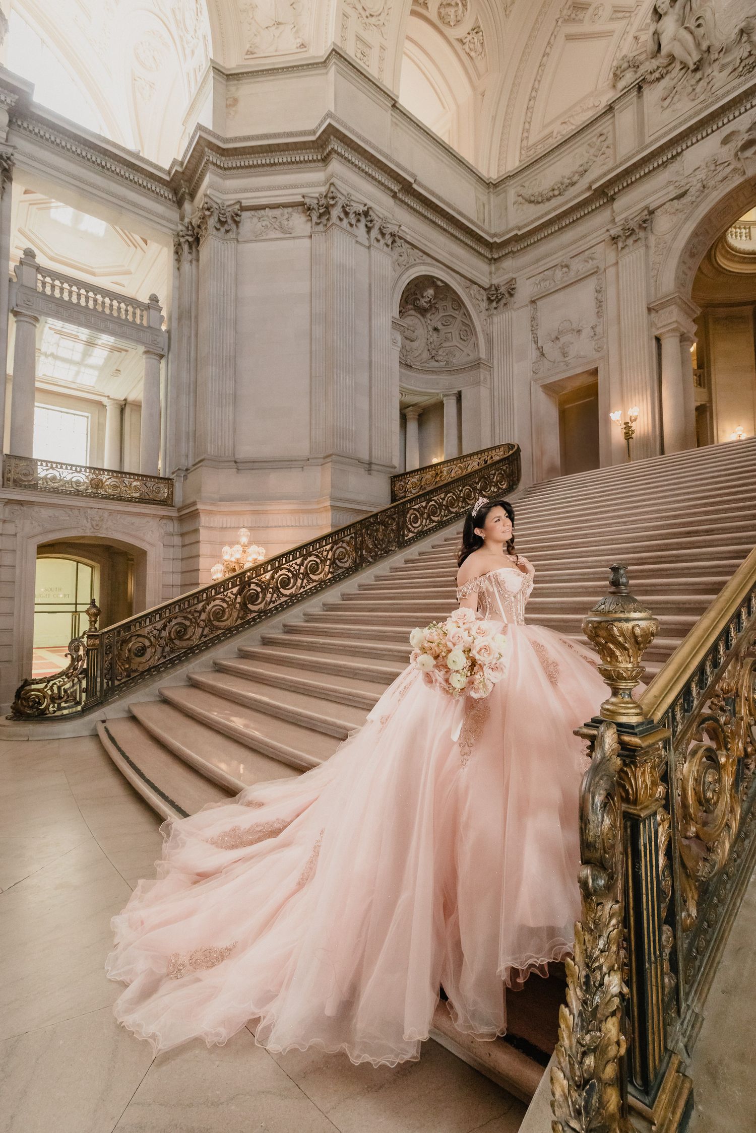 Quinceanera in blush dress poses on ornate staircase with intricate gold railings and classical architecture.