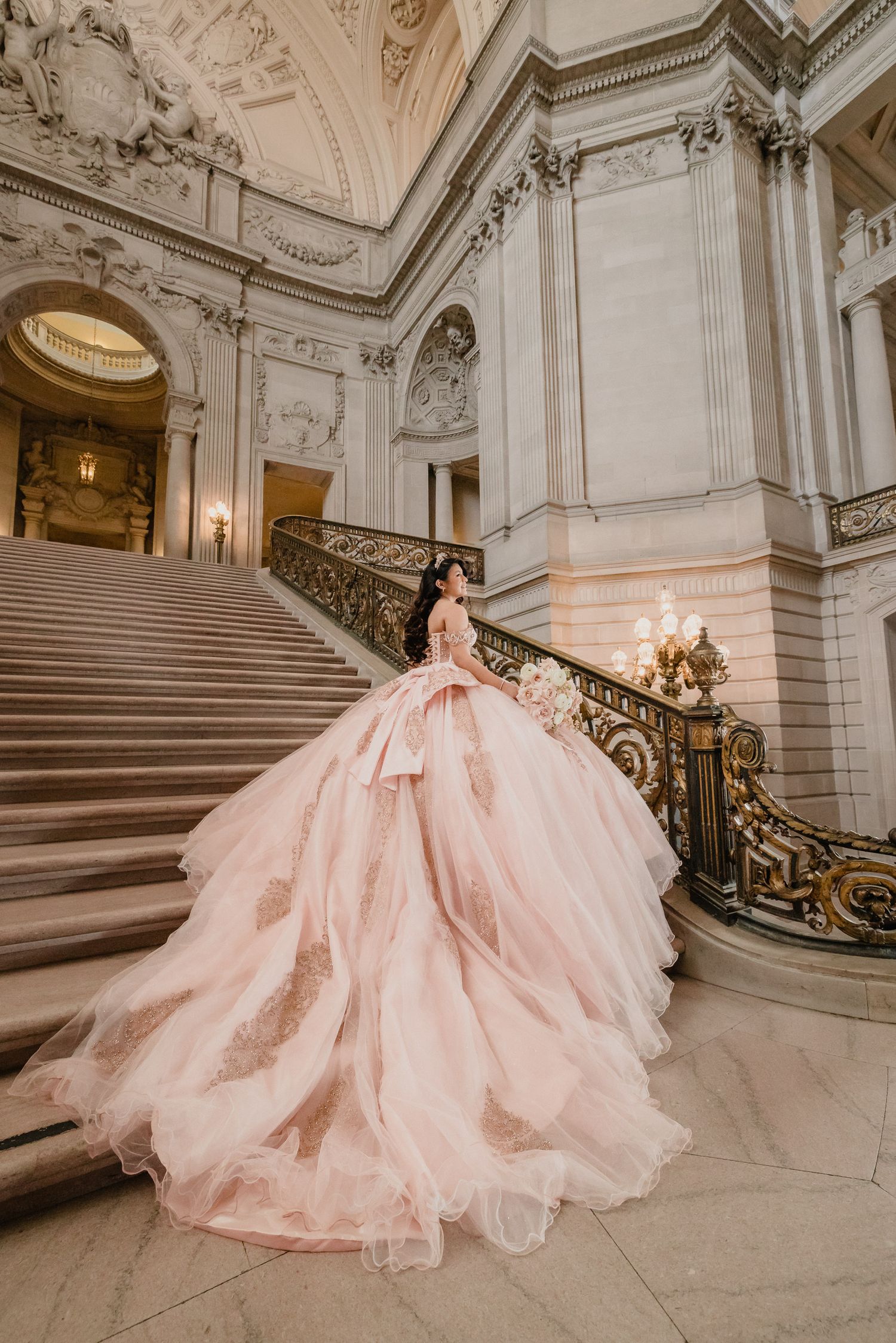Stunning quinceanera in pink dress showcases dramatic train on marble staircase in SF city hall.