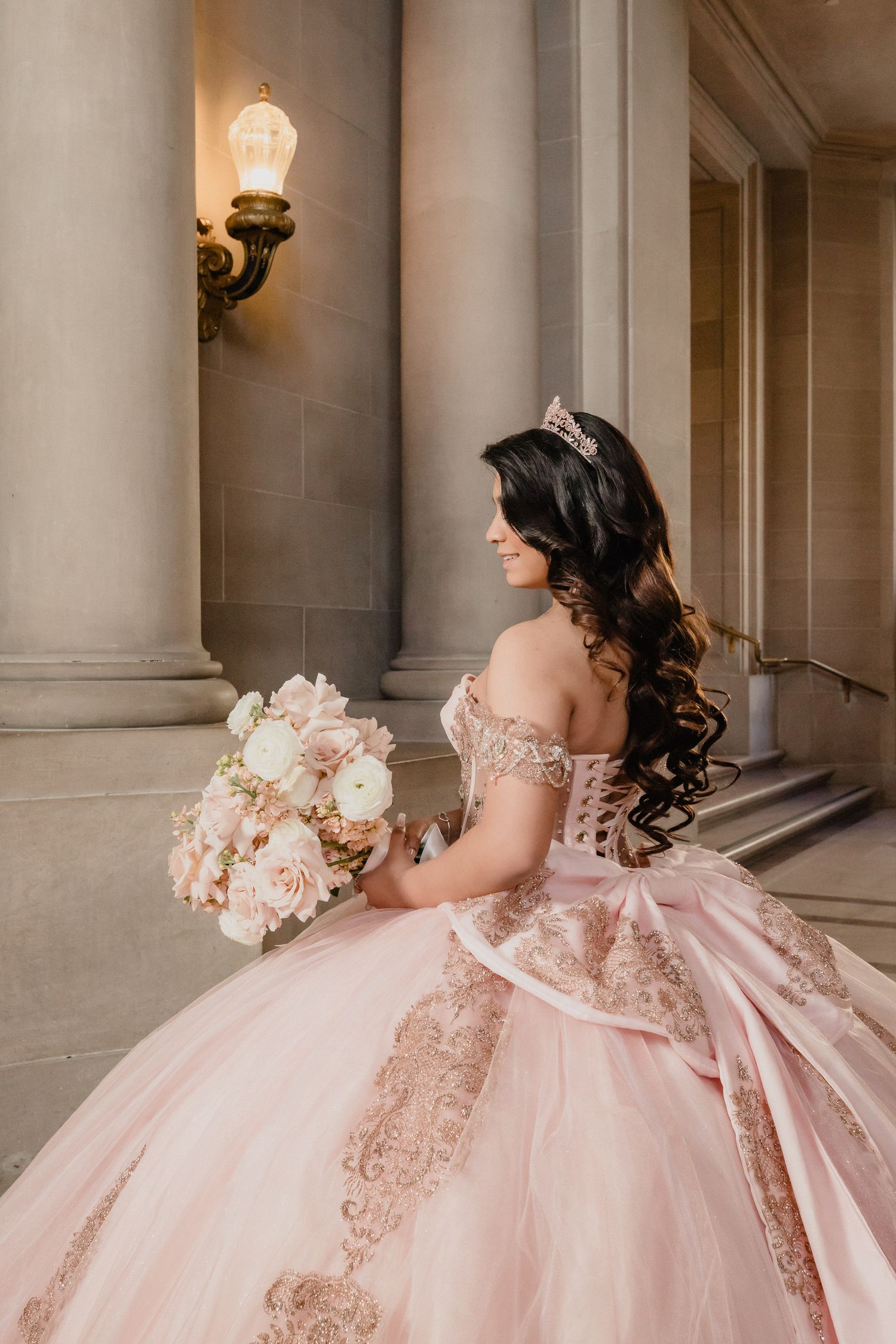 A quinceanera in an ornate blush pink quinceañera dress with lace details holds a bouquet in a grand columned hallway.