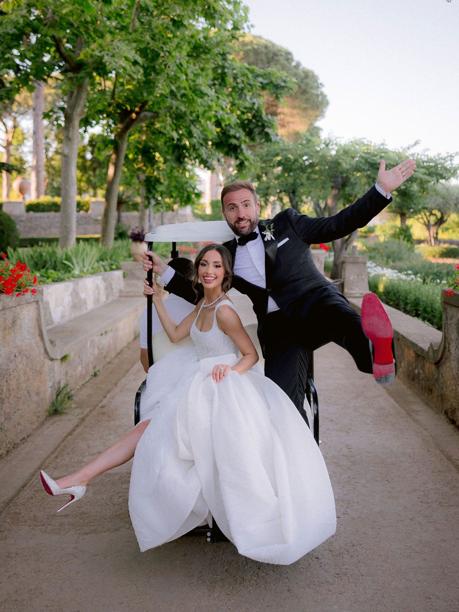 Newlyweds in formal attire strike playful poses on a garden pathway during their wedding photoshoot.